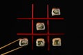 Tic-tac-toe game made with delicious sushi rolls and chopsticks on black background, flat lay