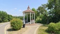 A tiled path leads to a metal gazebo with wooden benches, a bicycle lane and a urn next to it. Trees grow behind them. In the dist Royalty Free Stock Photo