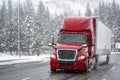 Tilted on the bend of the road red long hauler big rig semi truck with dry van semi trailer running on highway with forest and Royalty Free Stock Photo