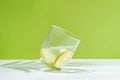 A tilted transparent glass of mineral water with lime on a green background lit by harsh daylight and a shadow from a plant.