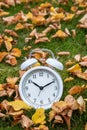 Time change, classic white alarm clock outside on grass and moss with fall color in many yellow birch leaves