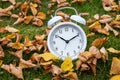 Time change, classic white alarm clock outside on grass and moss with fall color in yellow birch leaves
