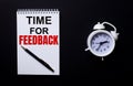 TIME FOR FEEDBACK is written in a white notepad near a white alarm clock on a black background Royalty Free Stock Photo