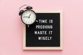 Time is precious waste it wisely. Motivational quote on letterboard and black alarm clock on pink background. Top view Flat lay