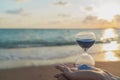 Sand glass in the hands symbolize the brevity of time. Background is seacoast and beautiful gold sundown