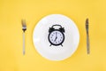 Time to eat. Breakfast time. View from above on white plate with alarm clock and knife and pork near on yellow background. 7 a.m Royalty Free Stock Photo