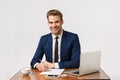 Time to make money. Good-looking office manager sitting his desk, writing report and smiling answering client, holding Royalty Free Stock Photo