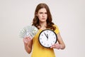 Time to make money! Strict teenager girl in yellow T-shirt holding big clock and dollars banknotes, looking at camera serious