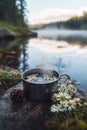 A tin camping cup filled with chamomile tea rests on a flat river stone. Steam rises in sync with distant mist over a lake, Royalty Free Stock Photo