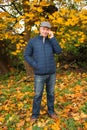 Tinted photo of an elderly man in a gray cap and blue jacket, who is standing in an autumn park among maple trees, is phoning and Royalty Free Stock Photo