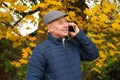 Tinted photo of an old man in a gray cap and a blue jacket, who is standing in the autumn park, is phoning and smiling, the Royalty Free Stock Photo