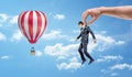 A tiny businessman held by a giant hand and hanging among blue sky near a striped air-balloon.