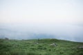 Tiny Human walks through mountainous area against background of field of grass and clouds.