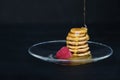 Tiny pancake cereal - trendy quarantine food. Pyramid of mini pancakes in plate with fresh raspberry and honey on black background Royalty Free Stock Photo