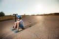 Tired active little caucasian boy in sport blue helmet and sneakers sitting on his black scooter on hot summer asphalt road on Royalty Free Stock Photo