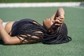 Tired African American lady athlete rests on green turf coat putting hand on forehead on sunny day