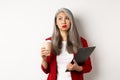 Tired asian female office worker holding clipboard and paper cup, drinking coffee and exhaling with exhausted face