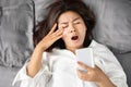 Tired Asian woman yawning in bed hand holding smart phone and rubbing her eyes , social addiction concept