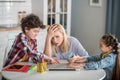 Tired blonde female sitting at table, holding her head, curly boy and dark-haired girl fighting over laptop