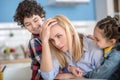 Tired blonde female sitting at table, holding her head, curly boy and dark-haired hugging her