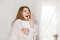 A tired, bored, beautiful girl holds a white pillow and yawns, covering her wide-open mouth with her hand. The girl with her eyes Royalty Free Stock Photo