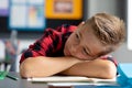 Tired, bored caucasian schoolboy at desk, resting head on crossed arms in class