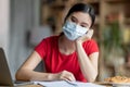 Tired boring adolescent asian girl in protective mask studies remotely in room interior