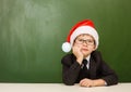 Tired boy in red christmas hat sitting near empty green blackboard Royalty Free Stock Photo