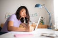 Tired chinese female college student doing homework writing on paper at home. Asian girl studying with laptop. Royalty Free Stock Photo