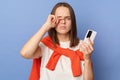 Tired dark haired woman wearing orange jumper over shoulders standing isolated over blue background using mobile phone long hours Royalty Free Stock Photo