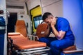 Tired doctor in uniform sits inside the ambulance car and puts his hands on head