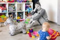 Tired of everyday household mother sitting on floor with hands on face. Kid playing in messy room. Scaterred toys and disorder.