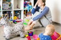 Tired of everyday household mother sitting on floor with hands on face. Kid playing in messy room. Scaterred toys and disorder.