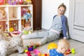 Tired of everyday household mother sitting on floor with hands on face. Kid playing in messy room. Scaterred toys and disorder.