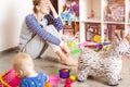 Tired of everyday household mother sitting on floor with hands on face. Kid playing in messy room. Scaterred toys and disorder.