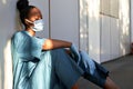 Tired exhausted woman african nurse wear face mask gloves sit on hospital floor. Royalty Free Stock Photo