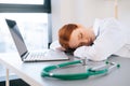 tired exhausted overworked young female doctor in white coat sleeping at desk with laptop and stethoscope on workplace. Royalty Free Stock Photo