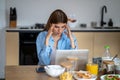 Tired exhausted workaholic woman with headache working on laptop sitting at table in kitchen.