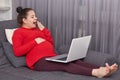 Tired expecting mother yawns as wants to sleep, works freelance at laptop computer, dressed in casual domestic clothes, sits at Royalty Free Stock Photo