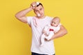Tired father with baby dummy dummy in his mouth, keeping hand on forehead, happy dad holding cute child in hands against yellow Royalty Free Stock Photo