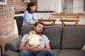 Tired Father With Baby Son Asleep On Sofa As Mother Works