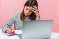 Tired female student leaning head on hand listening to lectures at laptop and making notes writing at notepad with pencil, Royalty Free Stock Photo