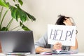 Tired frustrated woman sitting at table and holding card with word Help. Online working, learning from home