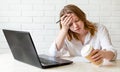 Tired girl holds his head with his hand and sitting at work desk at home studying at laptop with coffee Royalty Free Stock Photo