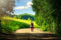 A Tired Guy in a Pink T-Shirt, Red Pants, and Bucket Hat, with Black Backpack and Long Pole Walks along Highland Country Road with Royalty Free Stock Photo