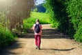 A Tired Guy in a Pink T-Shirt, Red Pants, and Bucket Hat, with Black Backpack and Long Pole Walks along Highland Country Road with Royalty Free Stock Photo