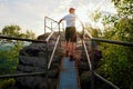 Tired hiker keep handrail on peak. Sunny spring daybreak in rocky mountains. Hiker with red baseball cap, dark pants and white Royalty Free Stock Photo