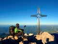 Tired hiker sit bellow crucifix on mountain peak. Iron cross at Alps mountain top. Royalty Free Stock Photo