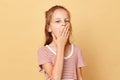 Tired little brown haired girl wearing striped t-shirt standing isolated over beige background covering mouth with palm yawning