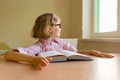 Tired little girl student looks out the window while sitting at her desk with a big book. School, education, knowledge and Royalty Free Stock Photo